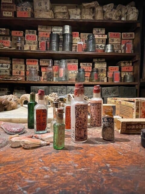 Tiny medicine glass bottles with Chinese writing on them. In the background is shelves of boxes with Chinese writing on them.
