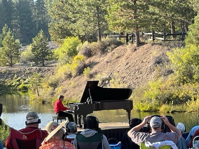 A large Steinway piano set against a mountain background with a young man playing it and audience members in the foreground.