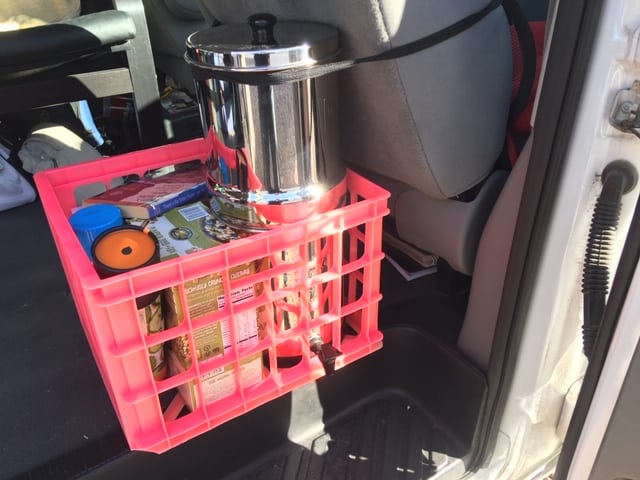 Red crate with a large silver cylindrical water filter system in it. It is secured to the back of a car's seat.